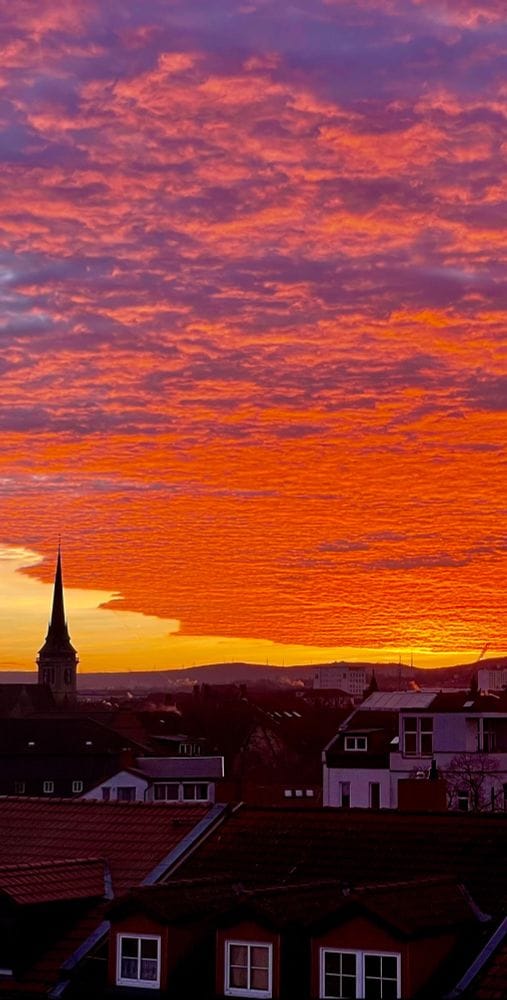 Die Sonne geht auf im Dezember 2023, alles ist in rote Farbe getaucht, man sieht eine Kirchturmspitze im linken unteren Bildbereich sowie Teile von Gründerzeithäusern. Die Wolken sehen wie Wellen aus. (C) Herr Montag.