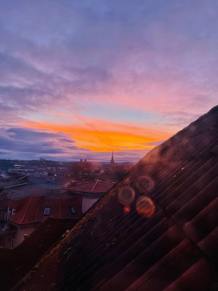 Sonnenaufgang über der Stadt, ringsherum sind Wolken und Dächer zu sehen. Ein wenig Morgenrot am Horizont. (C) Herr Montag.
