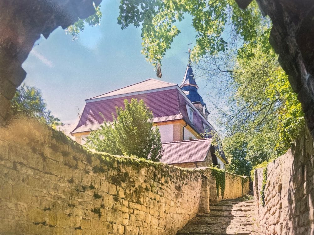 Eine alte Treppe führt hinauf, links sieht man ein Gebäude und ringsherum grüne Zweige von Büschen. Der Himmel ist blau, es ist sommerlich.