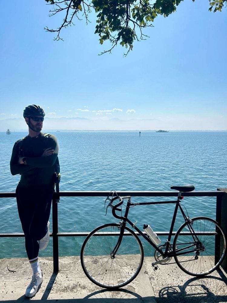 Herr Montag lehnt lasziv am Bodensee, man sieht ihn im Hintergrund. Das Wasser ist Azur. Neben ihm steht sein Vintage Rennrad.