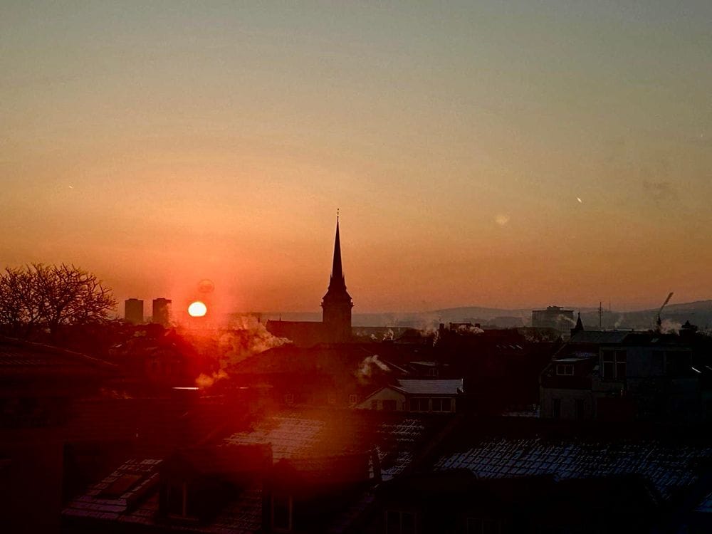 Sonnenaufgang über der Stadt: man kann scherenschnittartig die Umrisse erkennen, links eine Sonne, mittig eine Kirchturmspitze und in der Winterkälte dampfende Schornsteine (C) Herr Montag