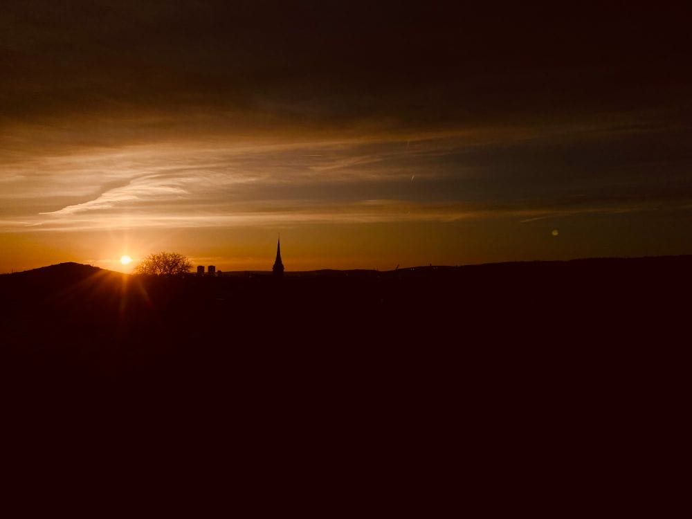 Sonnenaufgang über der Stadt, das Bild ist extrem dunkel gehalten, man sieht ganz viel schwarze Fläche und eine kleine Sonne, die leuchtet und ein wenig des Himmels freigibt. Das Bild verursacht eher Unbehagen, als dass es fröhlich wirkt, wie es einen Sonnenaufgang sonst tun würde. (C) Herr Montag