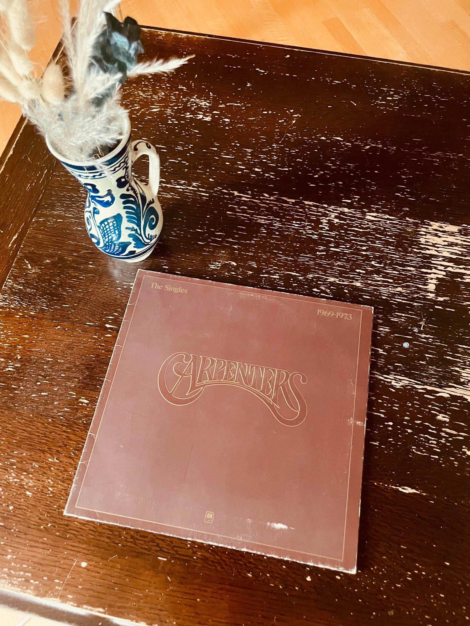 A vintage Carpenters album cover rests on a worn wooden table beside a decorative vase filled with dried grasses, creating a nostalgic and rustic vibe.