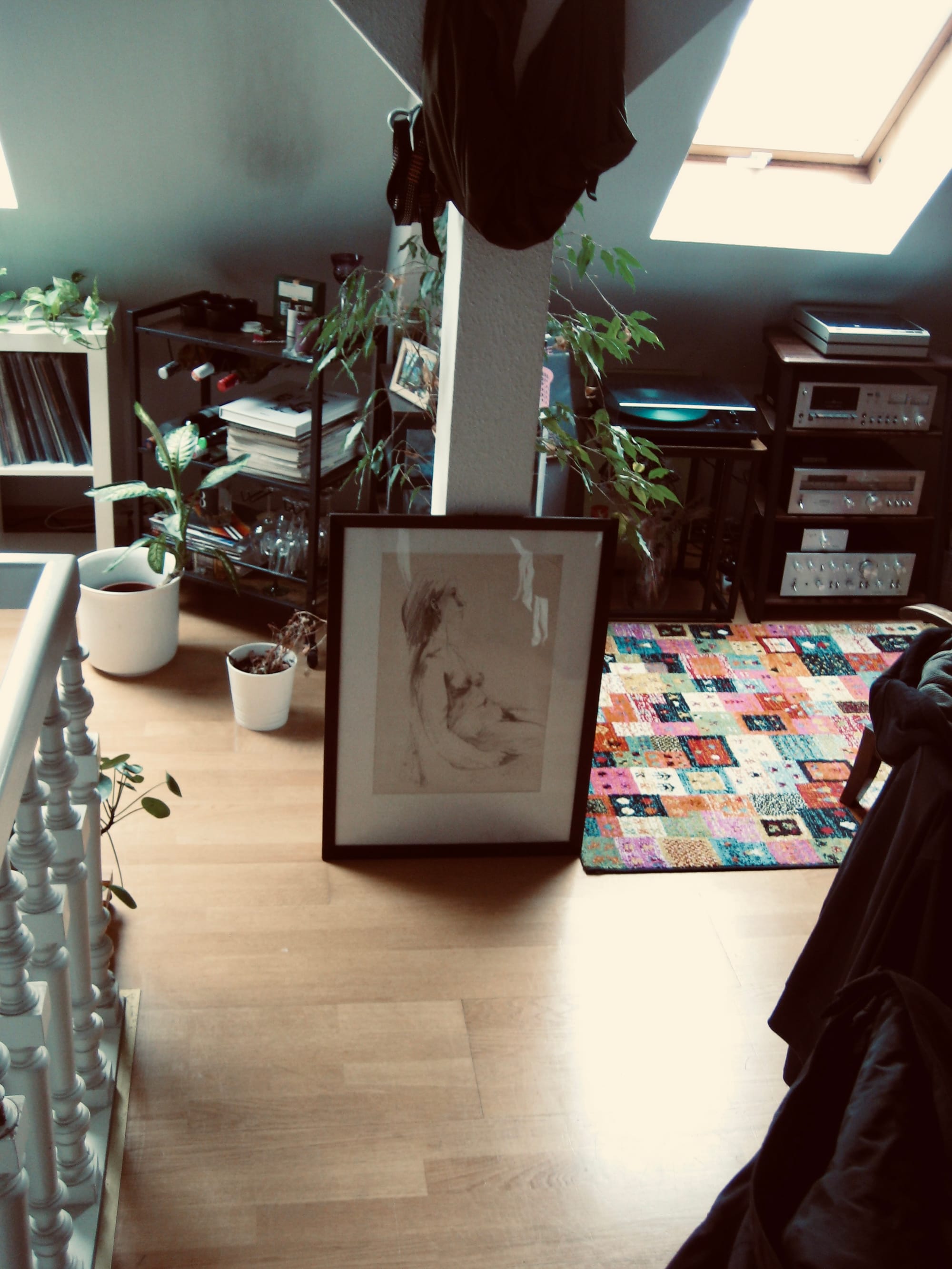 Das Bild "Nackte Frau" lehnt an einem Balken, daneben ein bunter Teppich und ein paar Pflanzen