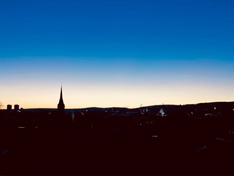 Sonnenaufgang über der Stadt, eine Kirchturmspitze ist zu sehen, im Winter geht die Sonne recht spät auf. (C) Herr Montag.